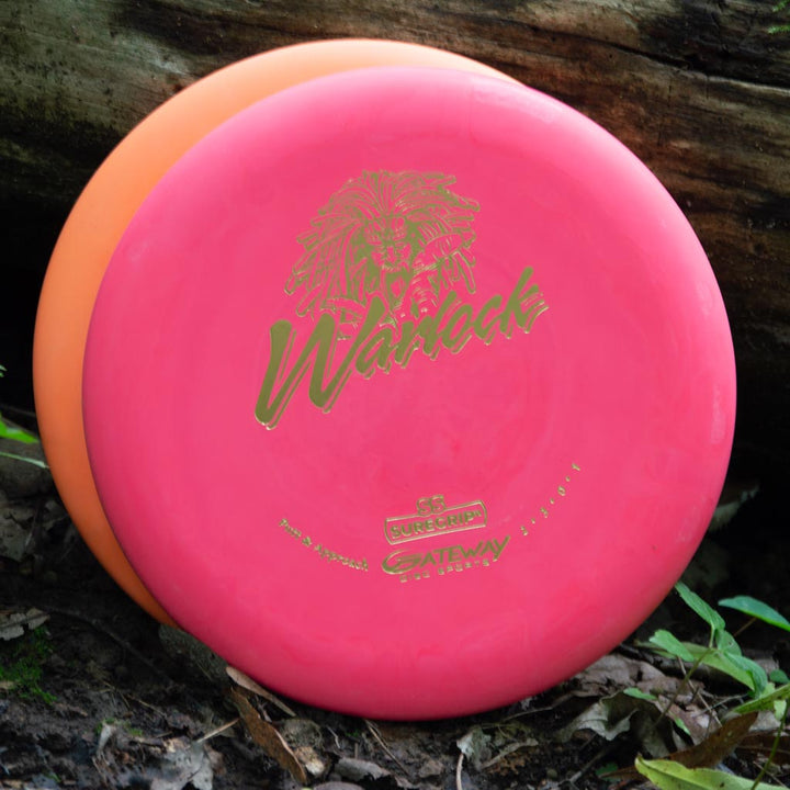 Two disc golf discs, one pink and one orange, lean against a log outdoors. The pink disc is a Warlock - Suregrip - SS putter featuring a striking lion graphic and "Warlock" in gold lettering.