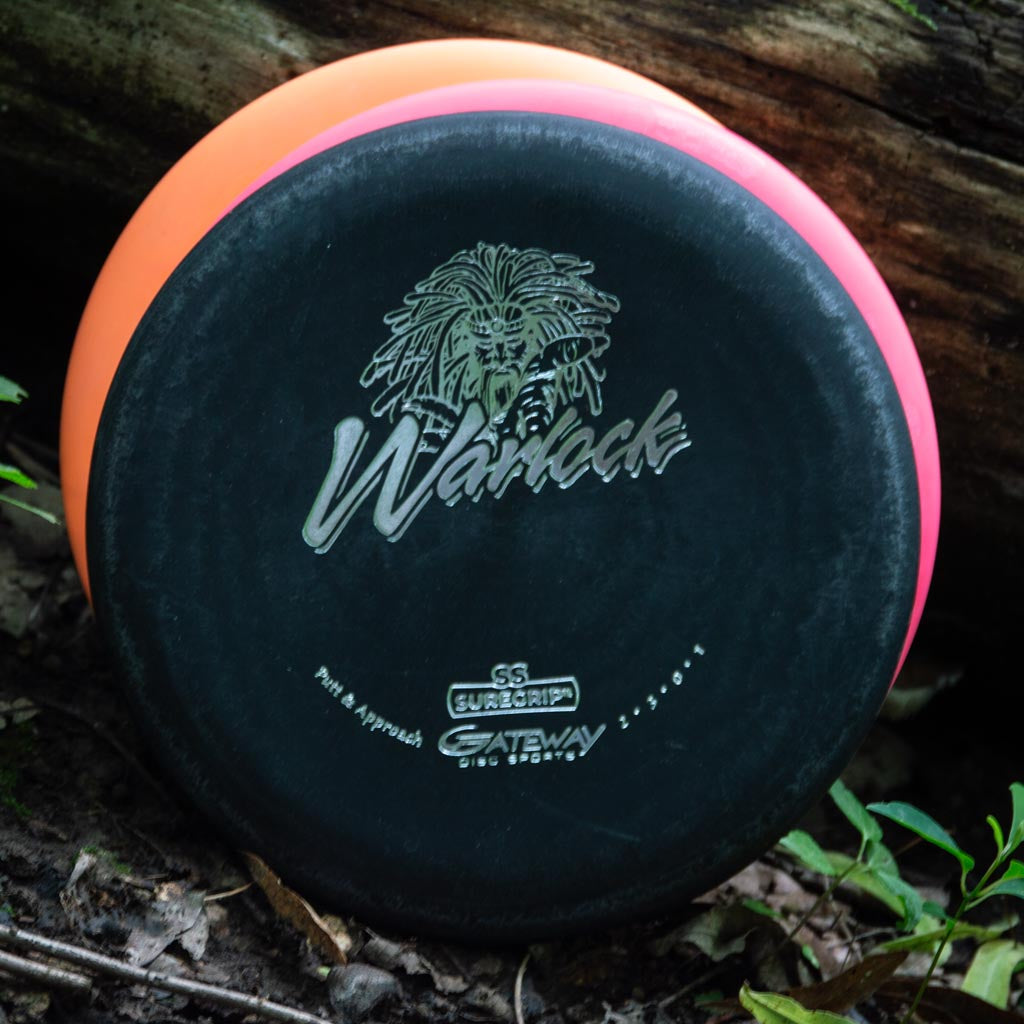 Two disc golf discs—a black Warlock - Suregrip - SS putter and an orange disc—lean against a log in the woods. The black one displays "Warlock" with a graphic and is recognized for its super soft Suregrip feel.