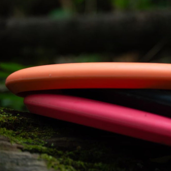 Close-up of two Warlock - Suregrip - SS disc golf putters, one orange and one pink, stacked on moss outdoors. The Super Soft (SS) plastic offers a unique feel ideal for precision shots.