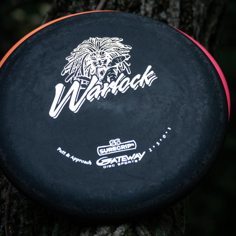 A black Warlock - Suregrip - SS putter with a warrior face graphic and "Gateway Disc Sports" logos rests against a tree.