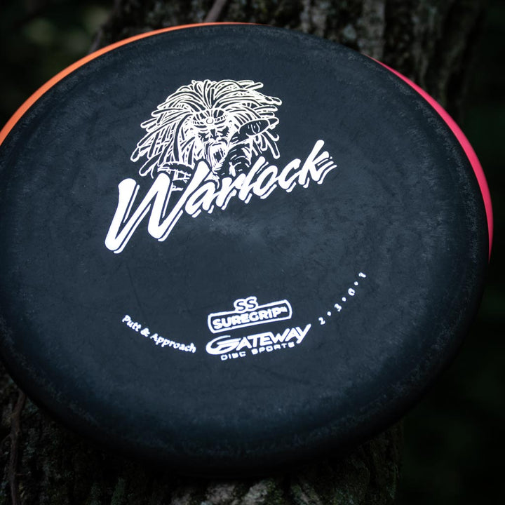 A black Warlock - Suregrip - SS putter with a warrior face graphic and "Gateway Disc Sports" logos rests against a tree.
