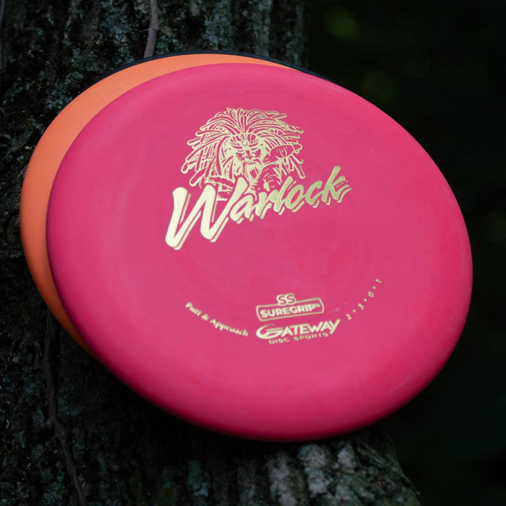 Two disc golf putters rest against a tree; the front disc is a pink Warlock - Suregrip - SS with gold logo and text, while the disc behind it is orange.
