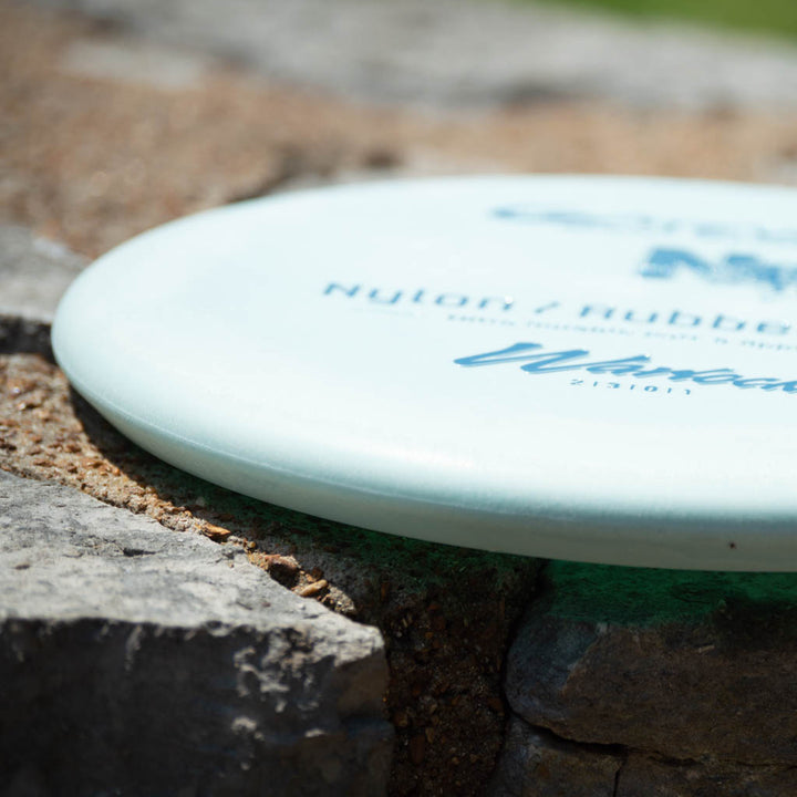 A close-up of a white Warlock - Nylon Superglow beadless putter with blue text, resting on rough stone outdoors.