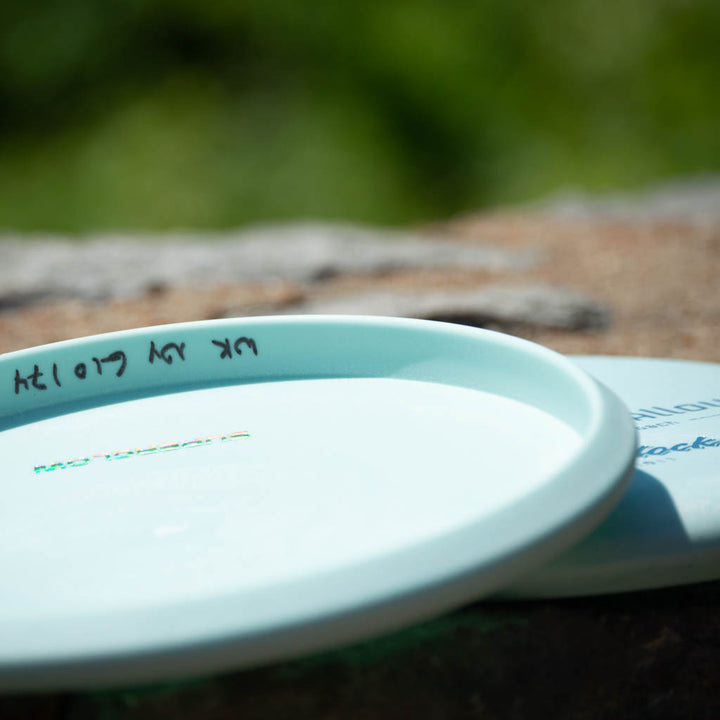 A pale blue Warlock - Nylon Superglow disc golf putter, known for its reliable beadless design and approach accuracy, sits on a rock with handwritten text on the rim against a blurred green background.