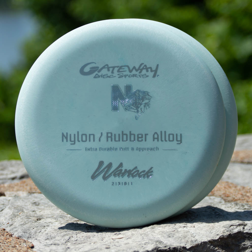 A light blue Gateway Disc Sports beadless putter labeled "Warlock - Nylon Superglow" rests on a rock outdoors, ready to enhance your approach game.