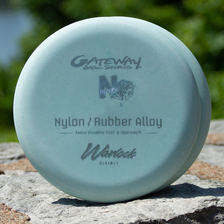 A light blue Gateway Disc Sports beadless putter labeled "Warlock - Nylon Superglow" rests on a rock outdoors, ready to enhance your approach game.