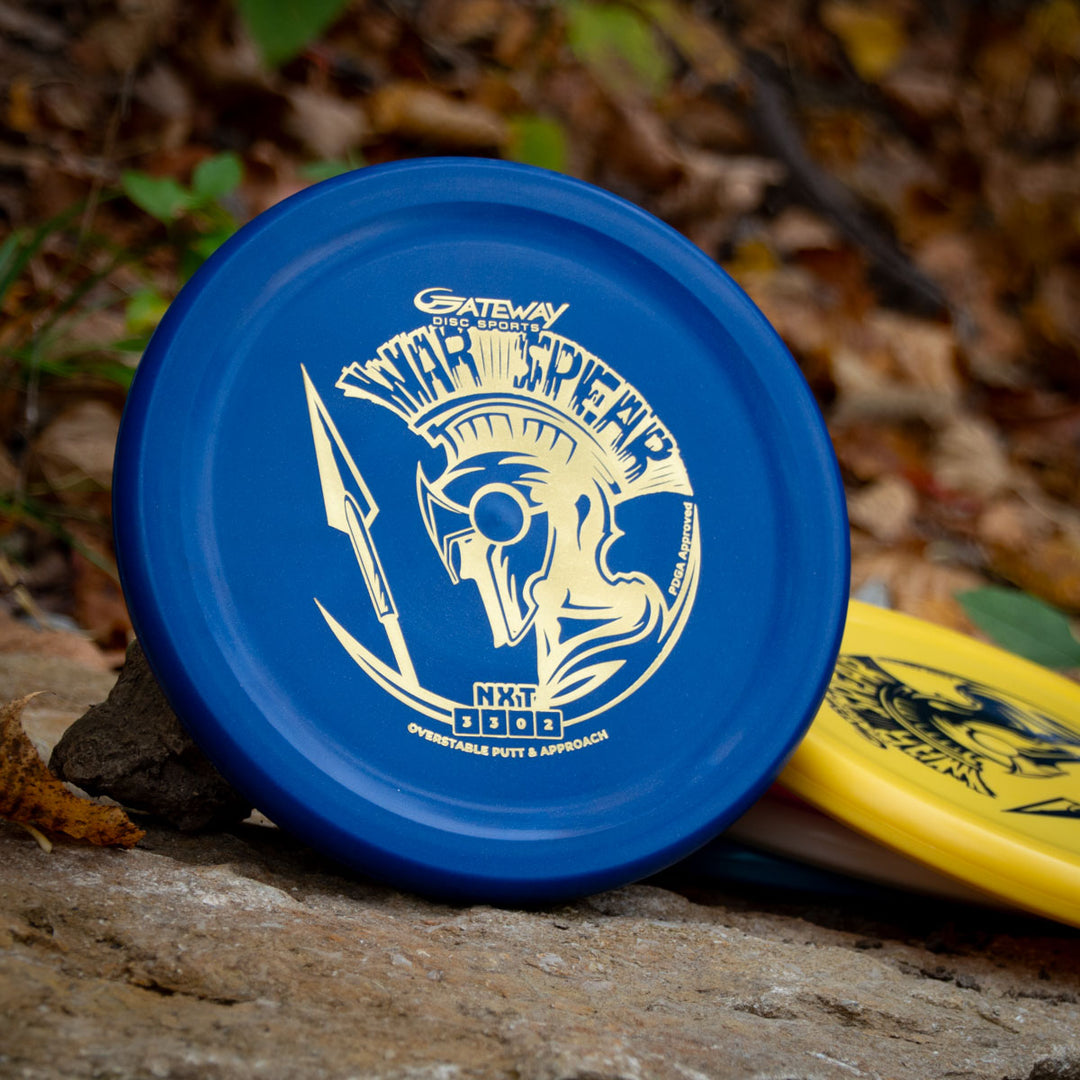 A blue Warspear - NXT disc golf putter, made from durable NXT plastic and featuring a gold Spartan helmet graphic, sits on a rock outdoors with a yellow disc in the background.