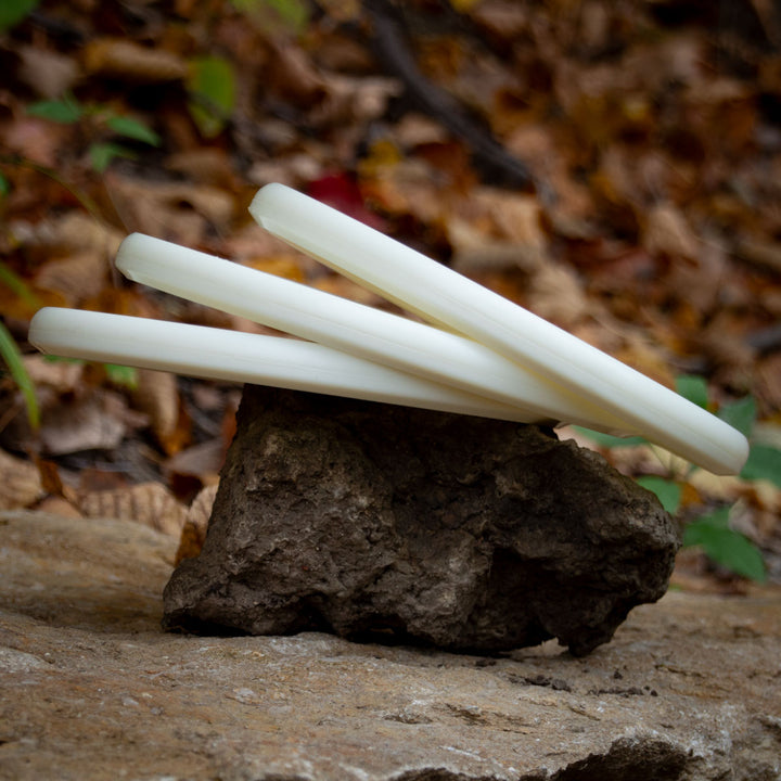 Three white Warspear - NXT Superglow cylindrical sticks are stacked on a rugged outdoor rock, with dry leaves and plants in the background.