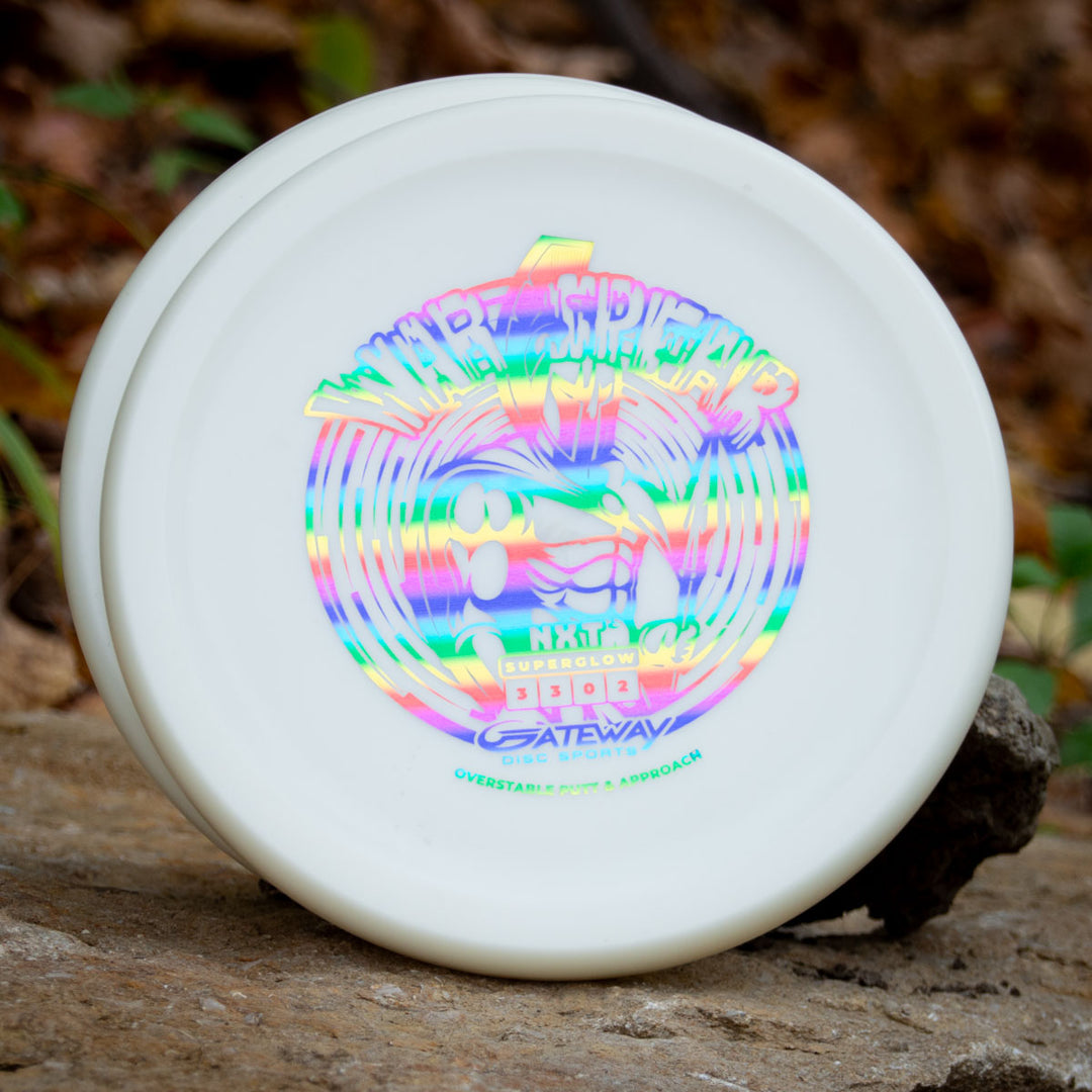 A white Warspear - NXT Superglow putt and approach disc with holographic artwork, Thumb Track design, and —Gateway Disc Sports—� branding rests on a rock outdoors.