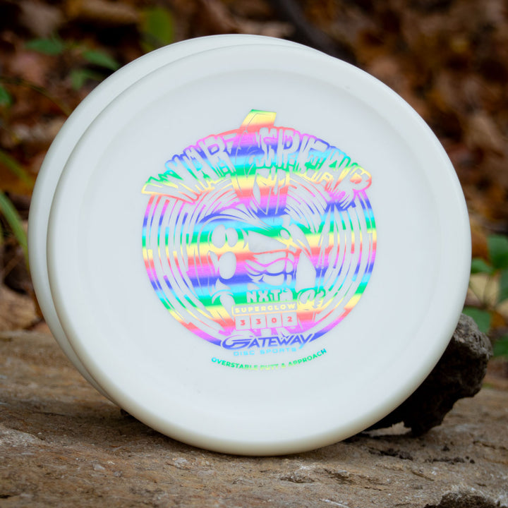 A white Warspear - NXT Superglow putt and approach disc with holographic artwork, Thumb Track design, and —Gateway Disc Sports—� branding rests on a rock outdoors.