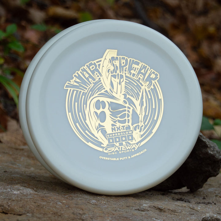 A white Warspear - NXT Superglow putt and approach disc, featuring a gold Spartan warrior design and the text "WARSPEAR, Gateway Disc Sports, Overstable Putt & Approach," rests on a rock outdoors.