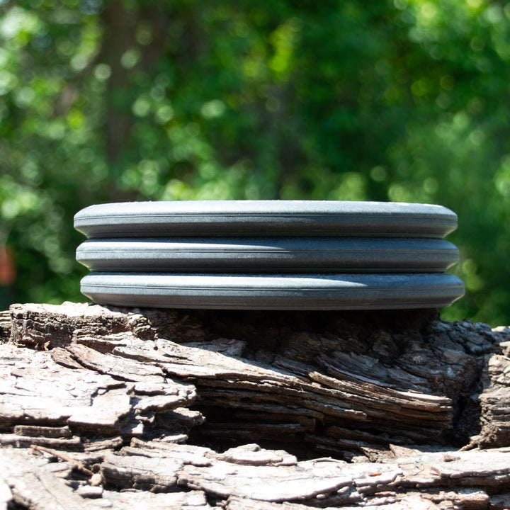 The Witch Doctor - Lunar, a black disc with three grooves, rests on textured wood outdoors, with blurred green foliage in the background.