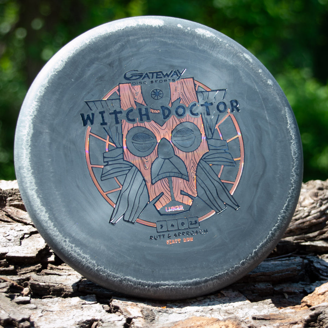 A Gateway "Witch Doctor - Lunar" gray beaded throwing putter with a tribal mask graphic rests on tree bark outdoors.