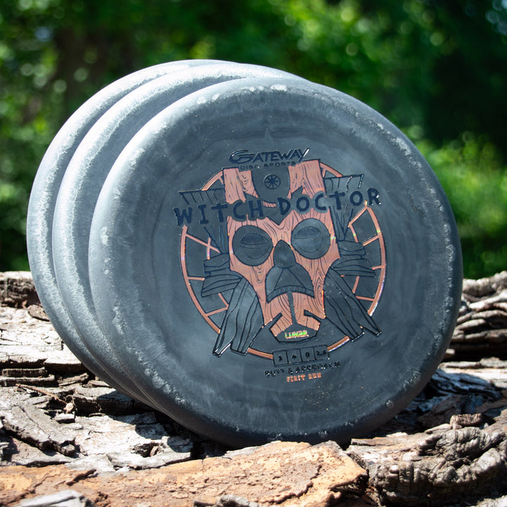 Three Witch Doctor - Lunar discs, each with a wooden mask design in black, are stacked on tree bark outdoors.