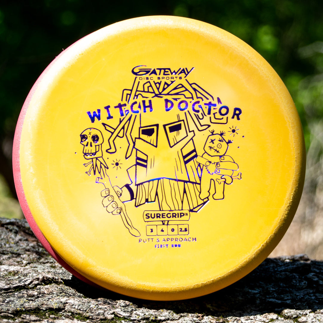 A yellow Gateway Disc Sports Witch Doctor SureGrip putter, featuring a mask, skull staff, and voodoo doll graphic, rests on a tree stump. 