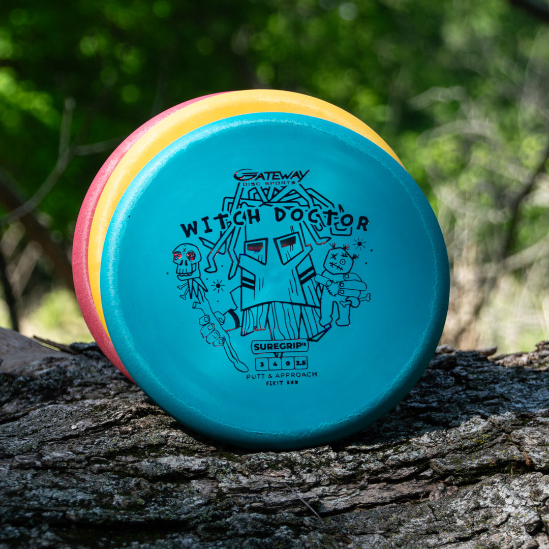 Three Witch Doctor - Suregrip - disc golf discs rest against a tree trunk. The front disc shows the Witch Doctor artwork, and their dependable Suregrip plastic stands out against a softly blurred forest background.