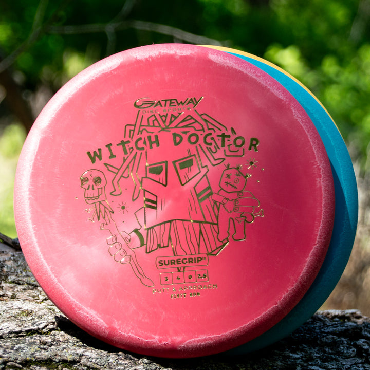 A Witch Doctor - Suregrip - disc golf putter in red with illustrated graphics rests on a log outdoors, offering reliable stability for your game, surrounded by lush green foliage.