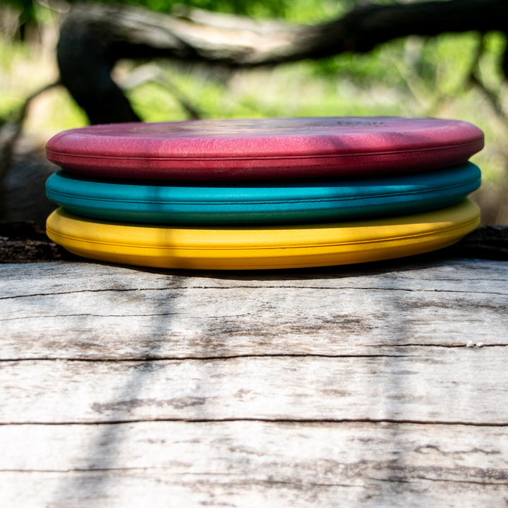 Three Witch Doctor - Suregrip - flying discs in red, teal, and yellow are stacked on a weathered wooden surface outdoors, with greenery in the background.