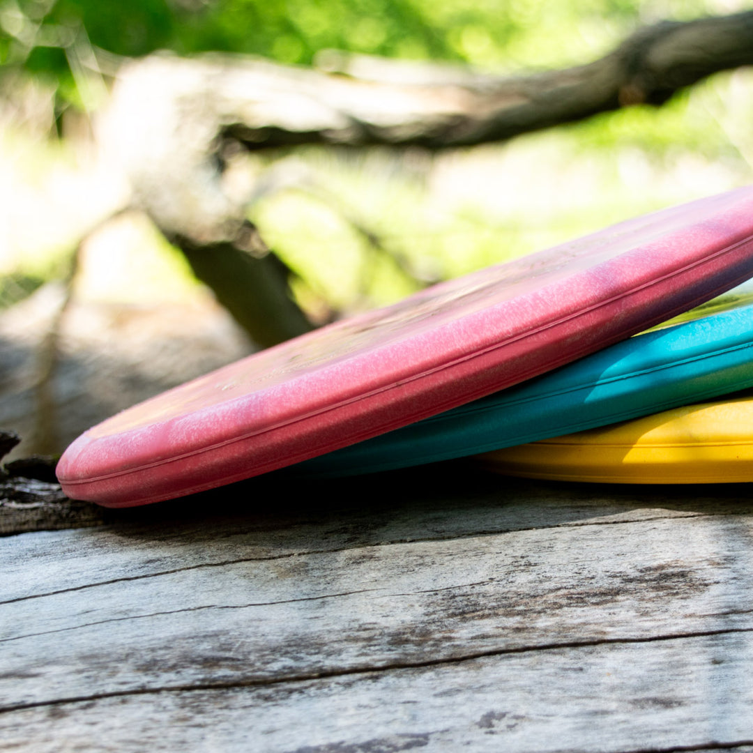 Three Witch Doctor - Suregrip - discs in red, blue, and yellow rest on a weathered wooden surface outdoors, highlighting their dependable stability and durable Suregrip plastic amid blurred greenery.