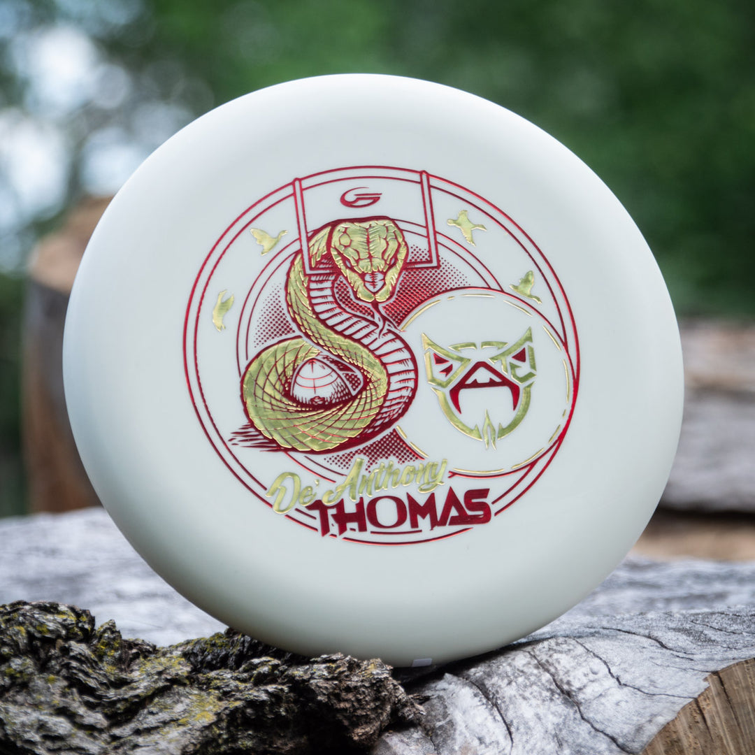 A white De'Anthony Thomas' Wizard - SS - Superglow disc with a red and yellow snake and face design rests on a stack of logs outdoors.