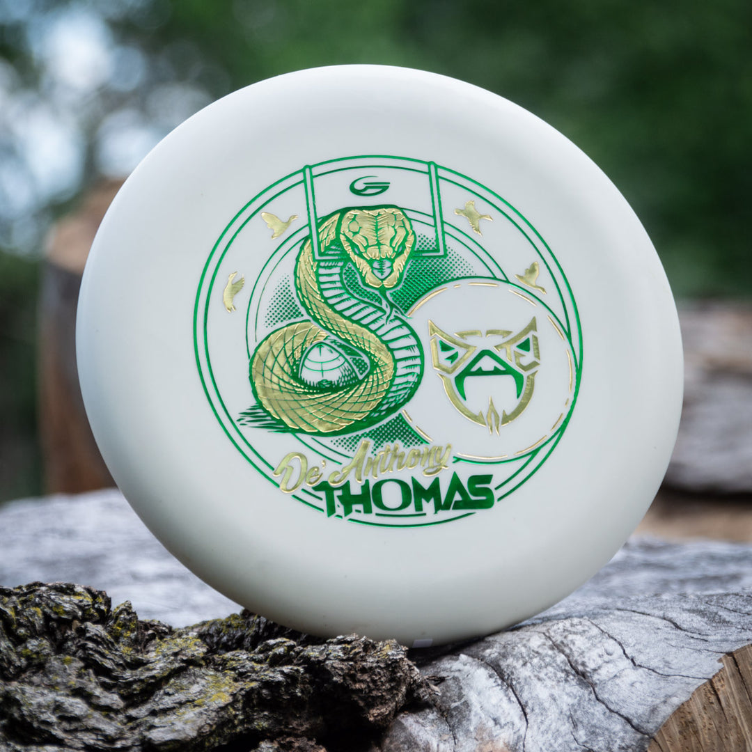 The De'Anthony Thomas' Wizard - SS - Superglow from Cardinal Disc Golf is a white disc with green and gold snake and mask artwork, propped against a log with a blurred outdoor background.