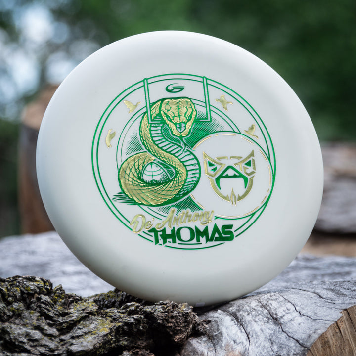 The De'Anthony Thomas' Wizard - SS - Superglow from Cardinal Disc Golf is a white disc with green and gold snake and mask artwork, propped against a log with a blurred outdoor background.
