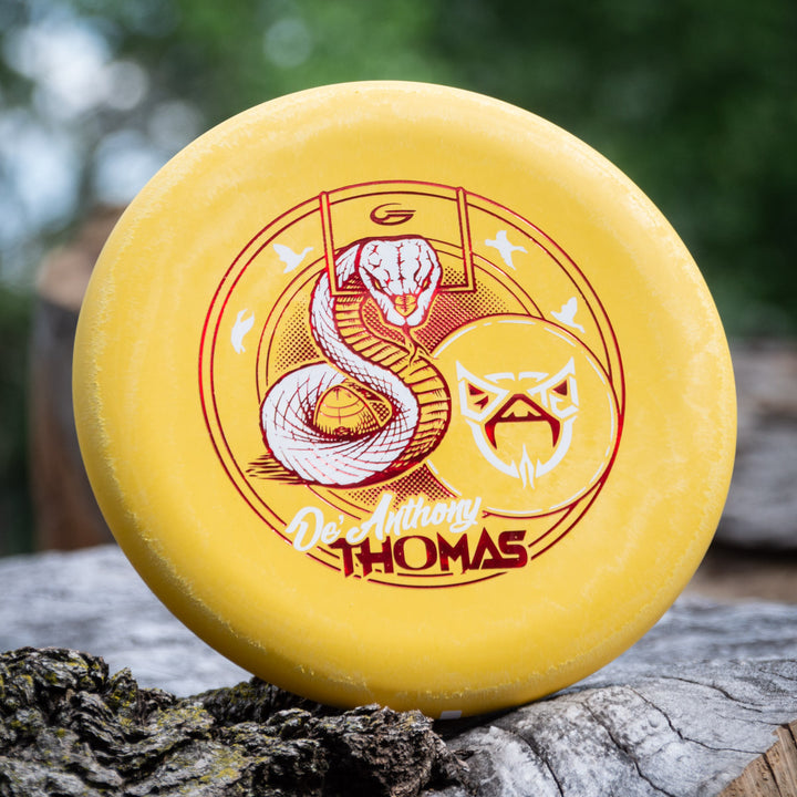 A yellow De'Anthony Thomas' Wizard - SS disc golf disc with a red snake and mask graphic is displayed outdoors on a tree trunk—ideal for fans of Black Mamba disc golf or De'Anthony Thomas memorabilia collectors.