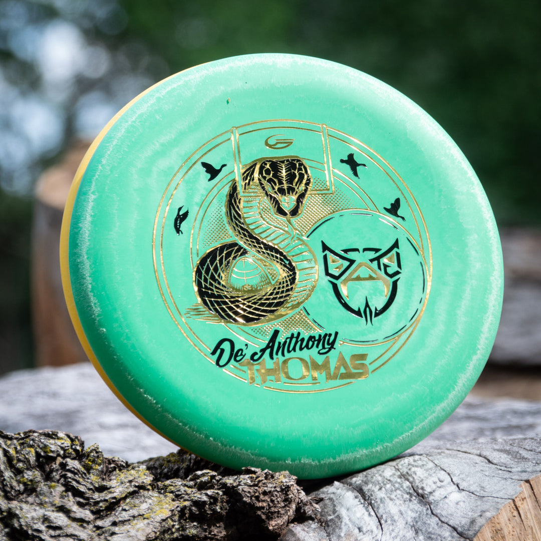 A green De'Anthony Thomas' Wizard - SS disc golf disc with gold snake, bird, and wolf artwork rests on a tree trunk outdoors.