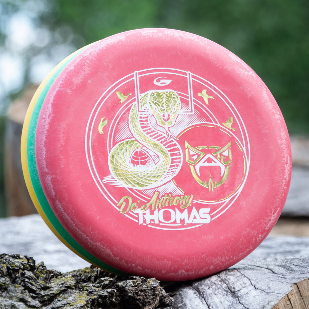 A red De'Anthony Thomas' Wizard - SS Disc Golf disc with a gold snake and bird design and "Devinciy Thomas" text rests on a tree trunk, set against a blurred green background.