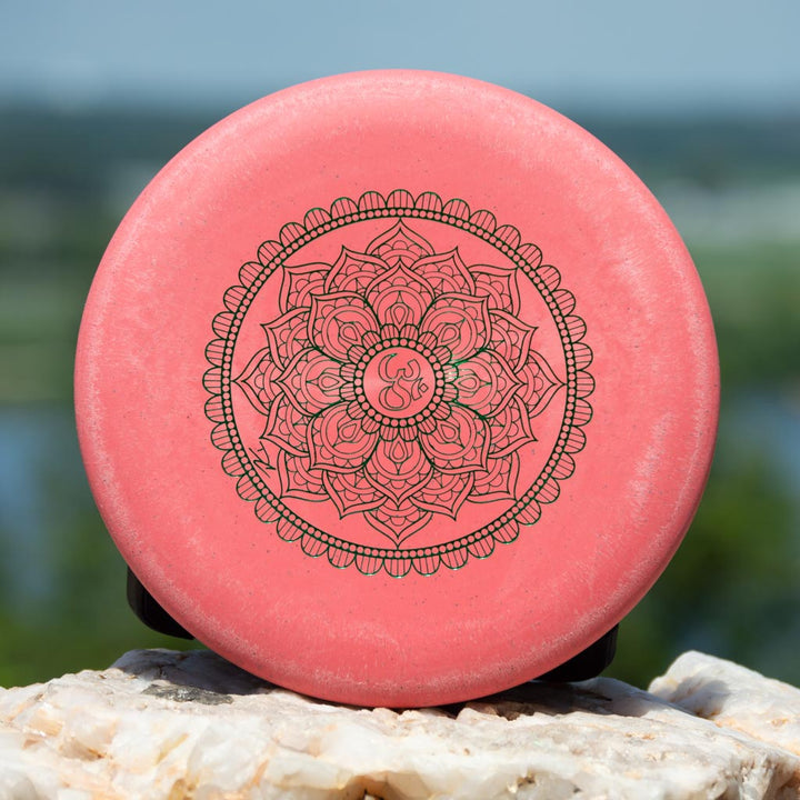 The Wizard - Mandala is a 160g pink disc golf disc featuring an intricate mandala pattern and Om symbol at its center. Flight numbers are printed along the rim. Displayed outdoors on a rock.
