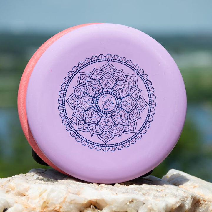 The Wizard - Mandala, a lavender disc golf disc with a dark purple mandala design and flight numbers, 160-Max weight, rests on a light-colored rock outdoors.