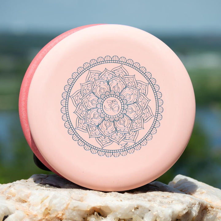 The Wizard - Mandala is a pink 160-Max weight disc golf disc featuring an intricate mandala design with an Om symbol at the center, shown resting on a rock. Add this unique disc to your bag and check its flight numbers for best performance.