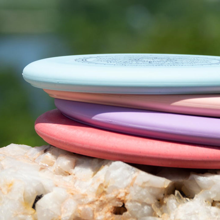 A Wizard - Mandala disc golf disc rests on a quartz rock, its vibrant colors and unique flight numbers standing out against a blurred green and blue outdoor backdrop.