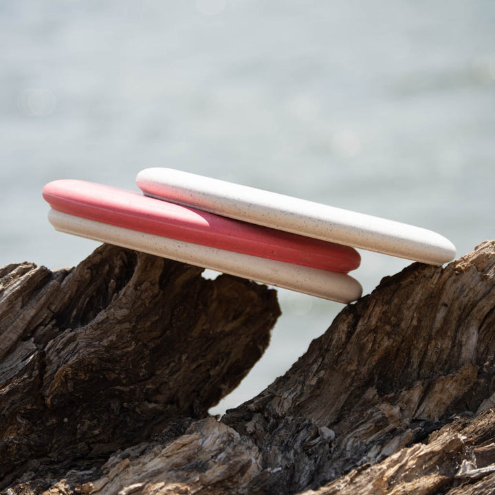 Two oval soap bars—one pink, one white—rest on driftwood with a blurred water backdrop, evoking the dependable performance of the Wizard - SSS - Organic Blend: a stable workhorse putter ready for mid-range drives in nature's fairway.