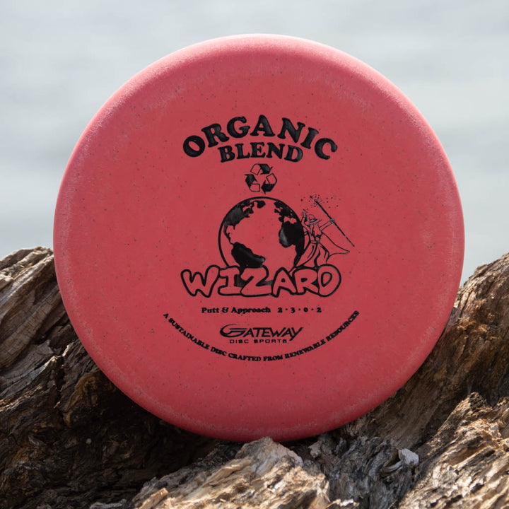 A red Wizard - SSSS - Hemp by Gateway Disc Sports, a stable workhorse putter, stands upright on wood with its logos and text clearly visible on the front.