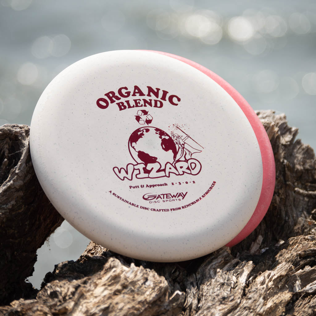 A white and pink Wizard - SSS - Organic Blend putter, known for stability and ideal for mid-range drives, with "Organic Blend Wizard" text, rests on driftwood by the water.