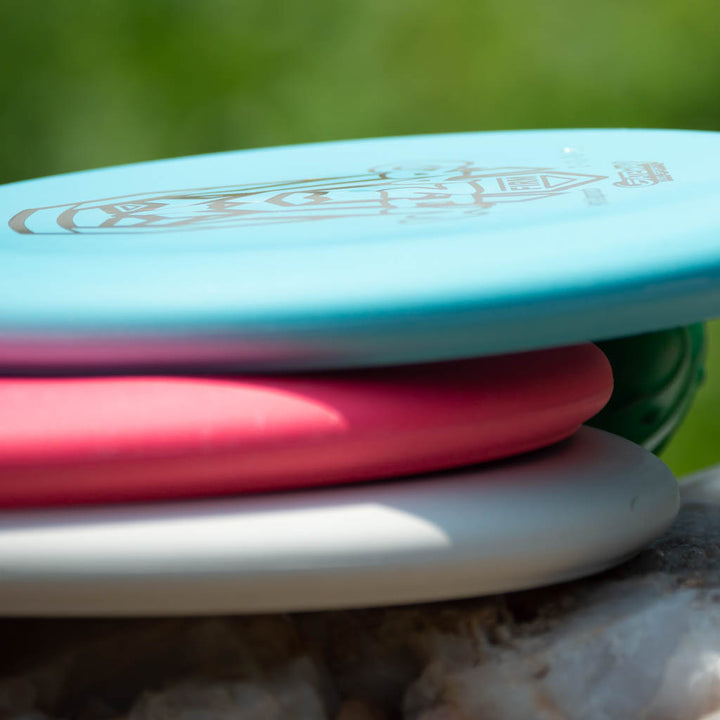 Three stacked Wizard - Firm putters in blue, pink, and white rest on a rock with a blurred green backdrop.