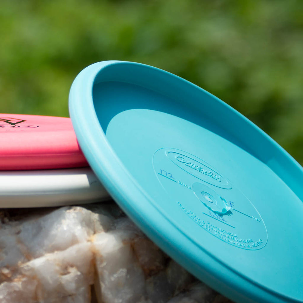 The Wizard - Firm disc golf discs—a blue, a red, and a white—rest on quartz with a blurred green backdrop, ready to be your stable workhorse even in windy conditions.