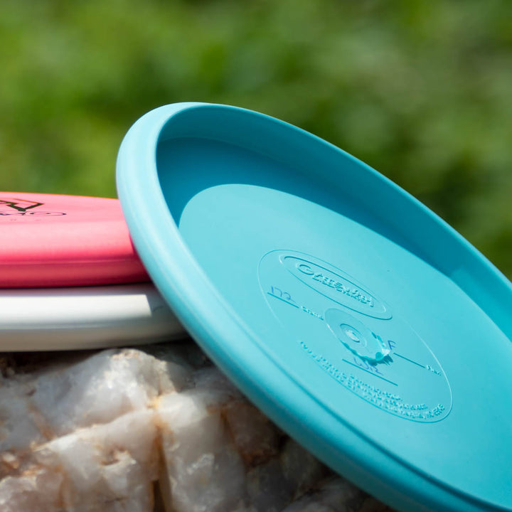 The Wizard - Firm disc golf discs—a blue, a red, and a white—rest on quartz with a blurred green backdrop, ready to be your stable workhorse even in windy conditions.