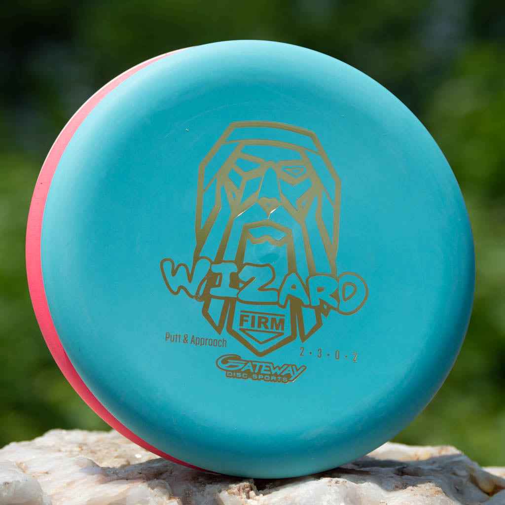 The Wizard - Firm putter features a blue and pink color, a gold wizard graphic, and "WIZARD FIRM" & "Gateway Disc Sports" text—ideal as your stable go-to disc, even in wind. It sits on a rock with a blurred green background.
