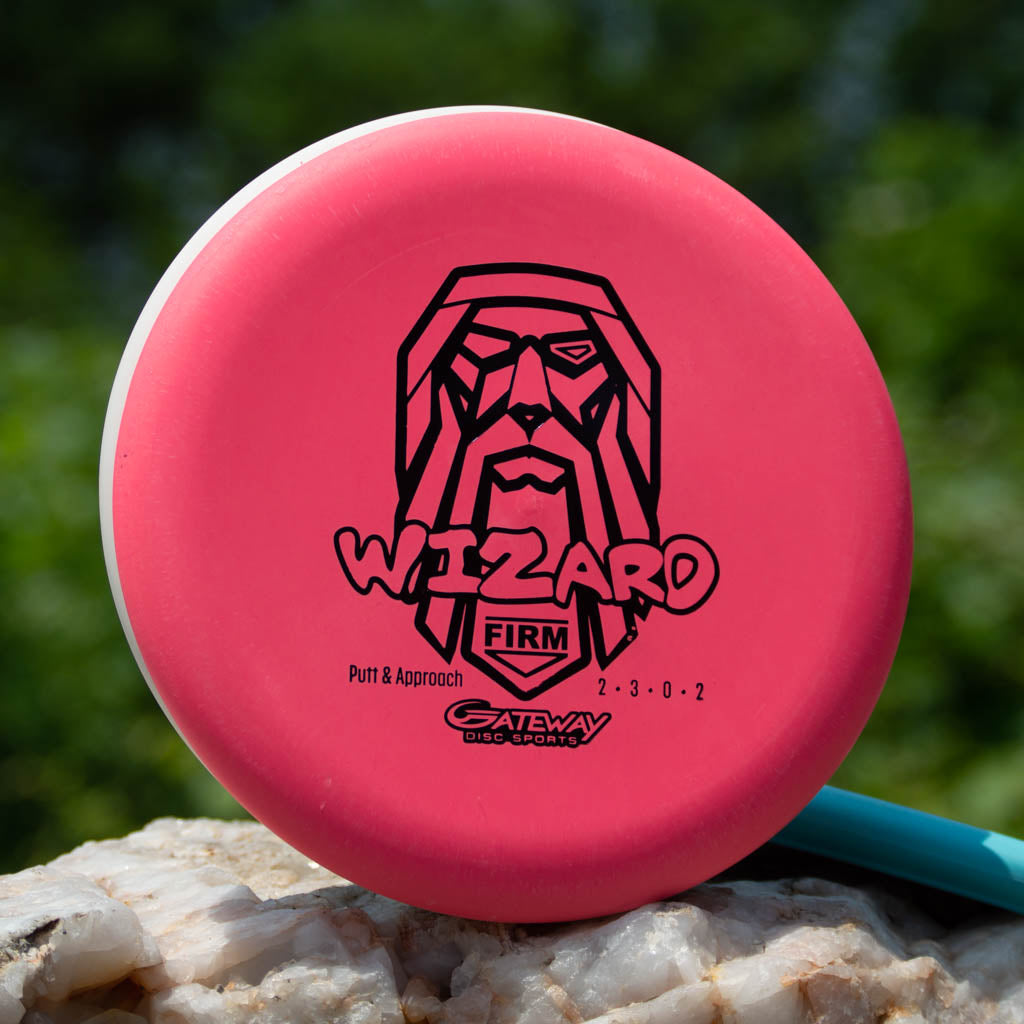 A pink Wizard - Firm putter from Gateway Disc Sports, a stable workhorse in wind, stands upright on a rock with a black wizard face logo against a blurred green background.