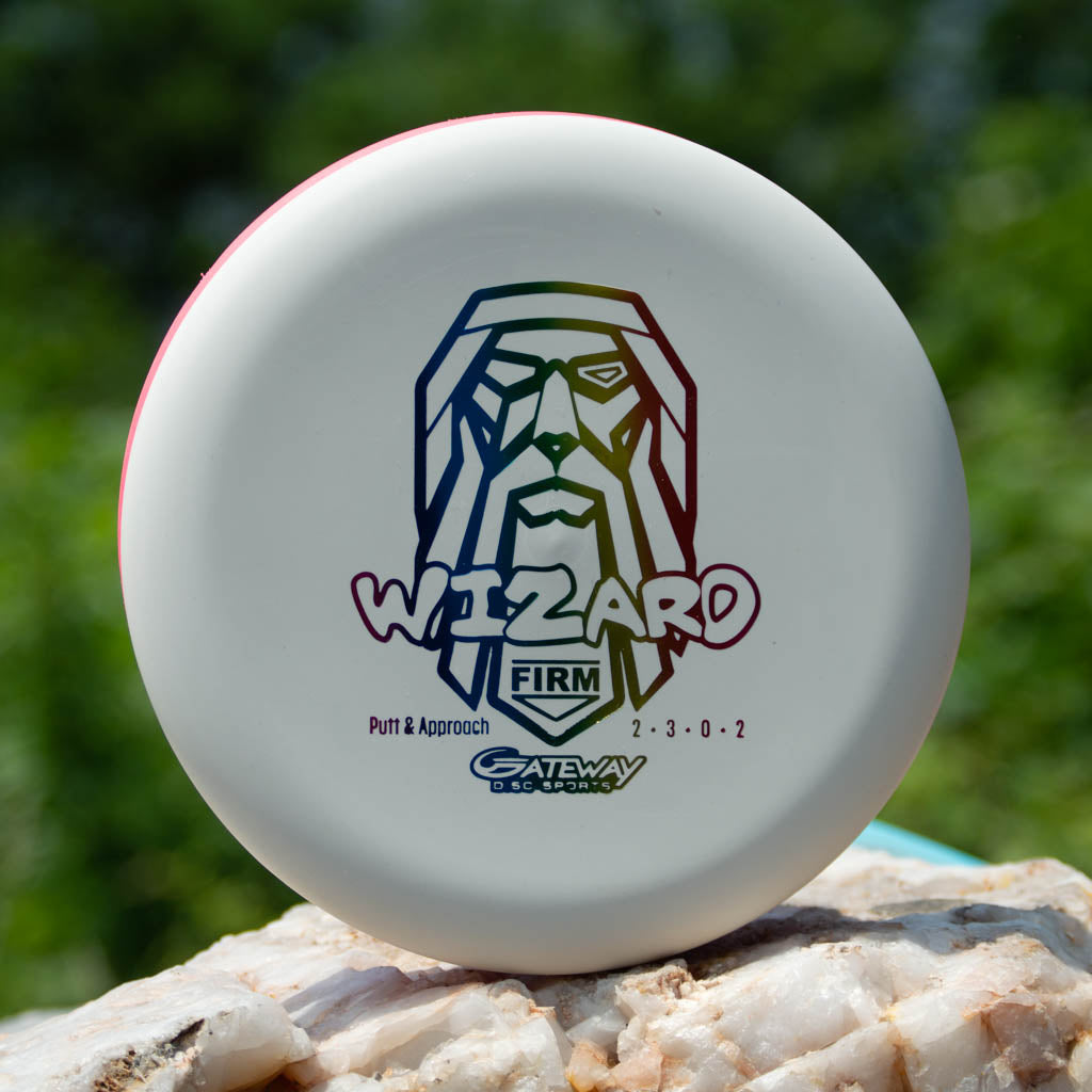 A white Wizard - Firm putter with a stylized wizard face graphic rests on a rock against a blurred green backdrop; this stable disc is an ideal workhorse for windy conditions.
