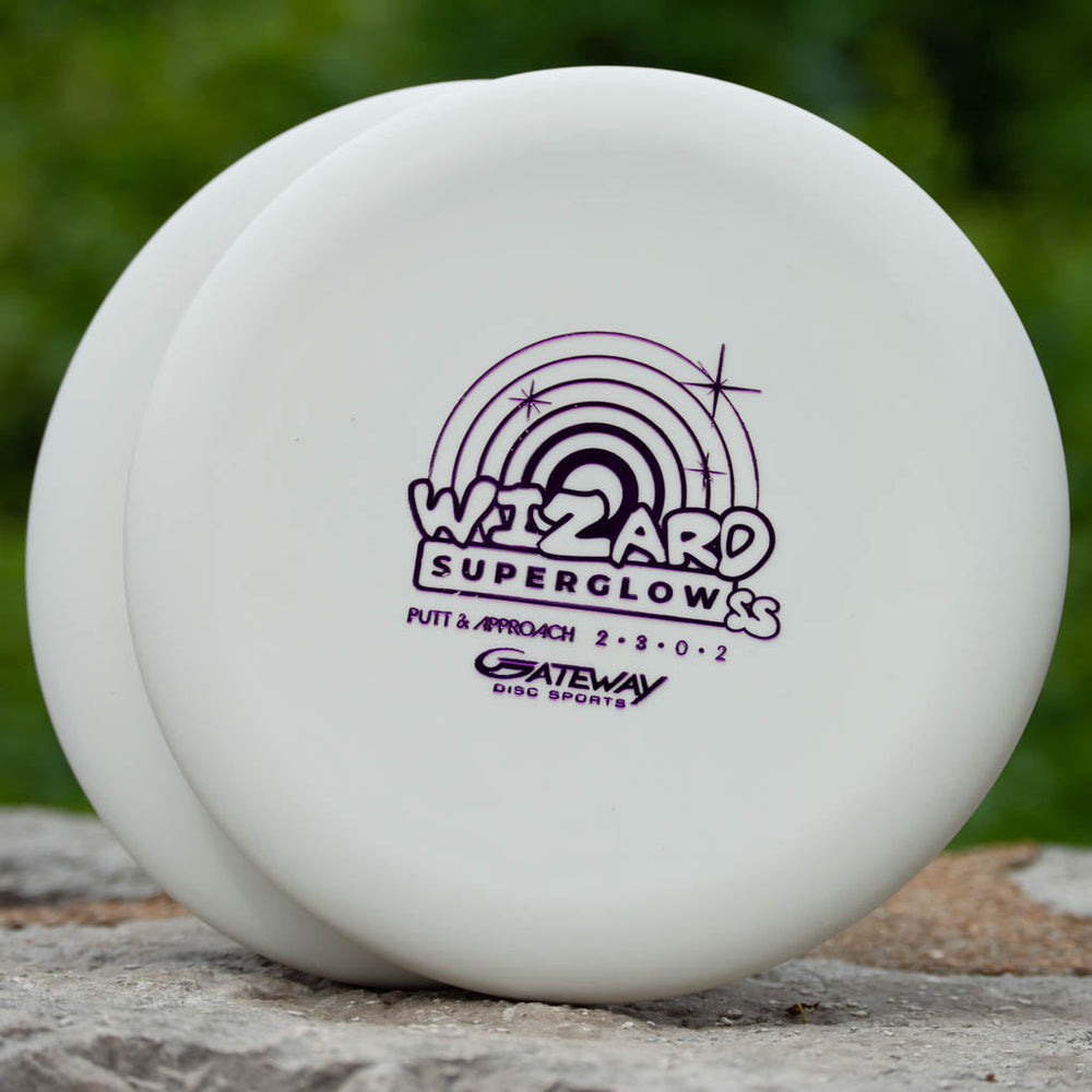 Two white Wizard - SS - Superglow disc golf putters with purple text and graphics rest on a rock amid greenery, highlighting this stable Gateway putter's performance in a natural setting.