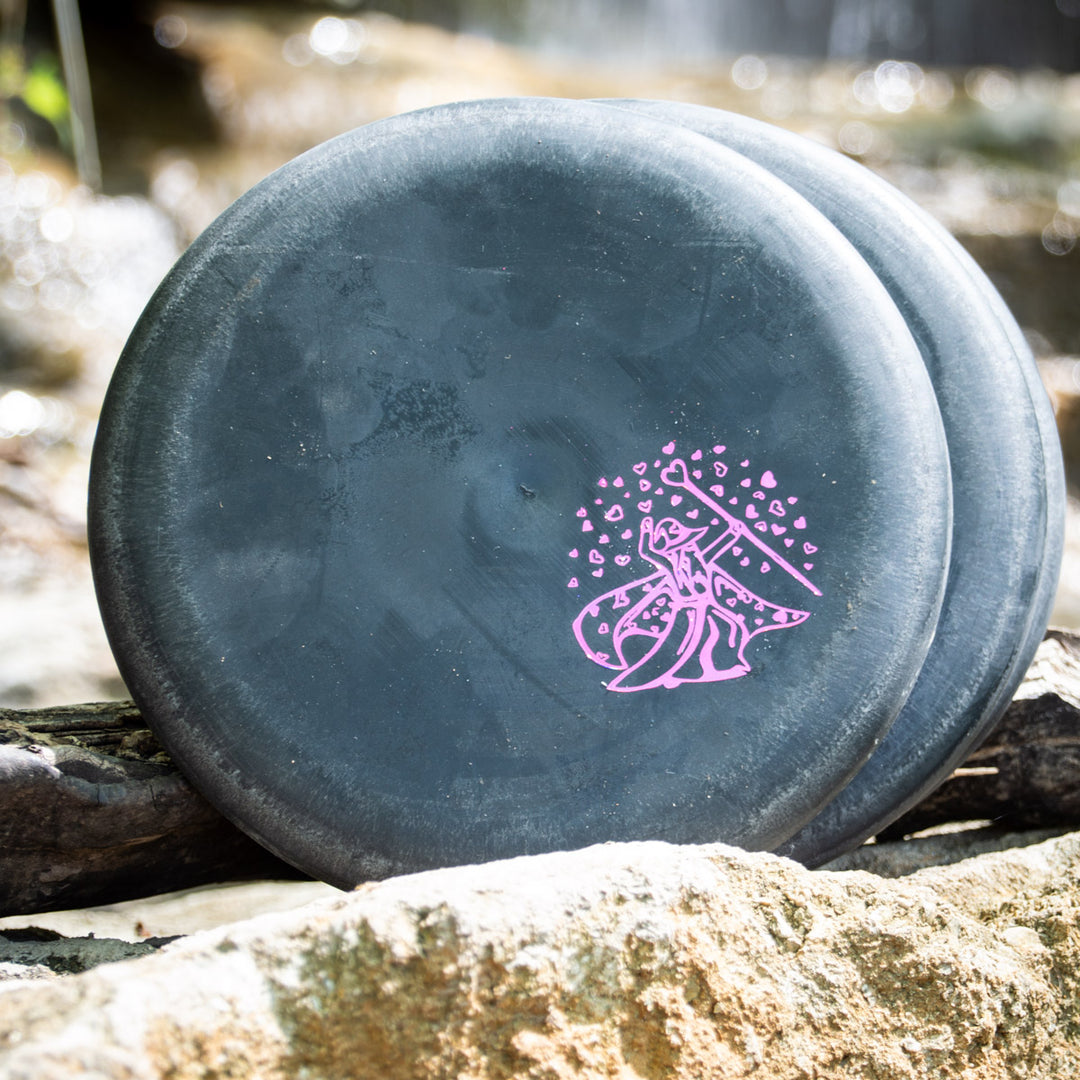 Three black Wizard SSS Hearts discs with pink graphics, recognized for consistency and control, are placed on rocks outdoors, with a blurred backdrop of water and foliage.