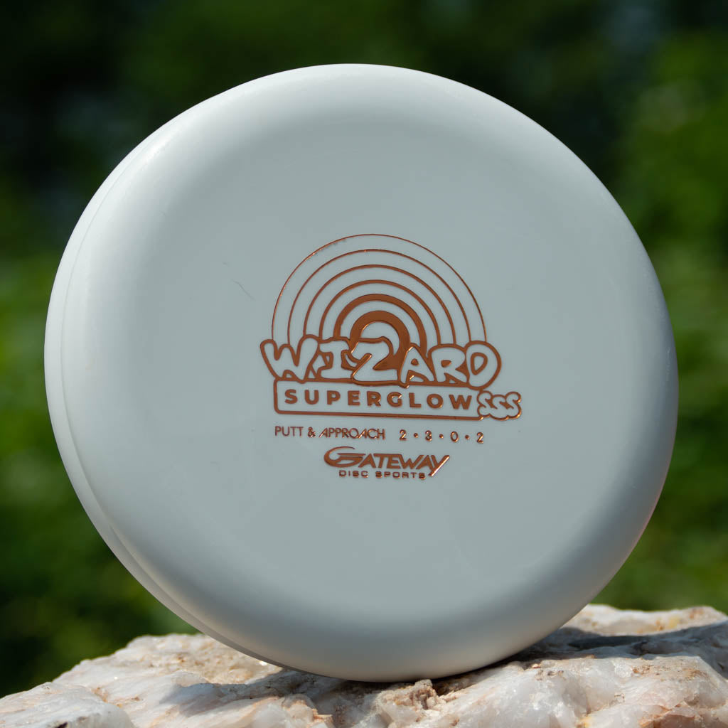 A white Wizard - SSS - Superglow putter from Gateway Disc Sports sits on a rock with greenery behind it, ready to be your stable go-to for putts and mid-range drives.