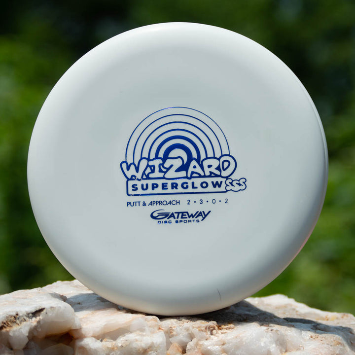 A white Wizard - NAKED - SSS - Superglow disc golf putter from Gateway Disc Sports, known for its stable performance on mid-range drives, sits on a rock outdoors.
