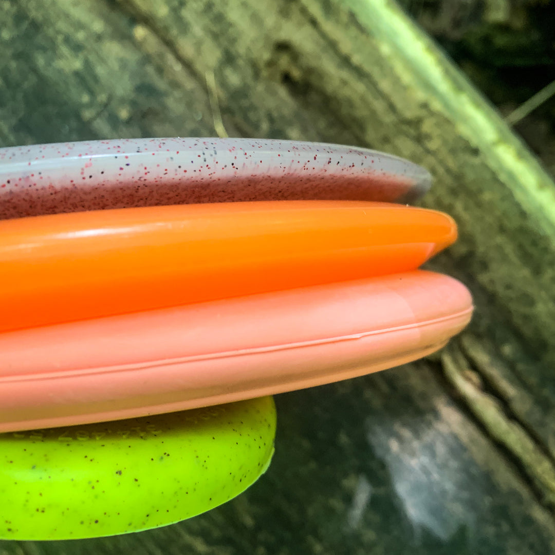 Close-up of four stacked discs from the Gateway Factory 2nd Mystery 55-Pack—gray, orange, pink, and green drivers and putters—with a blurred wooden surface in the background.