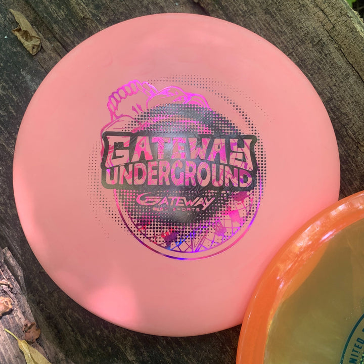 A pink disc from the Gateway Factory 2nd Mystery Promo Packs, featuring "Gateway Disc Sports" text, lies on a wooden surface beside a partially visible orange disc.