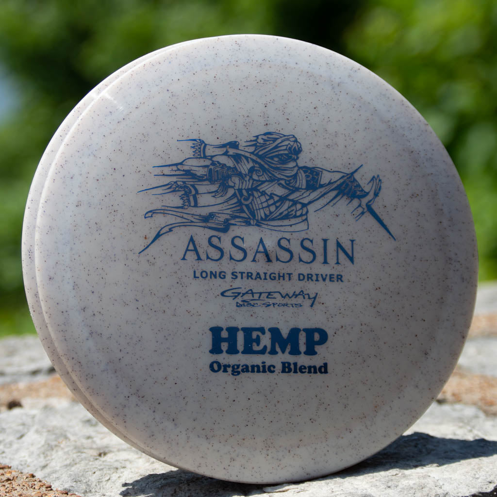 Close-up of a white Assassin - Diamond Hemp distance driver with "ASSASSIN," an archer graphic, "Diamond Hemp Blend," and "Gateway Disc Sports" text, resting on a rock. Features a neutral flight path.