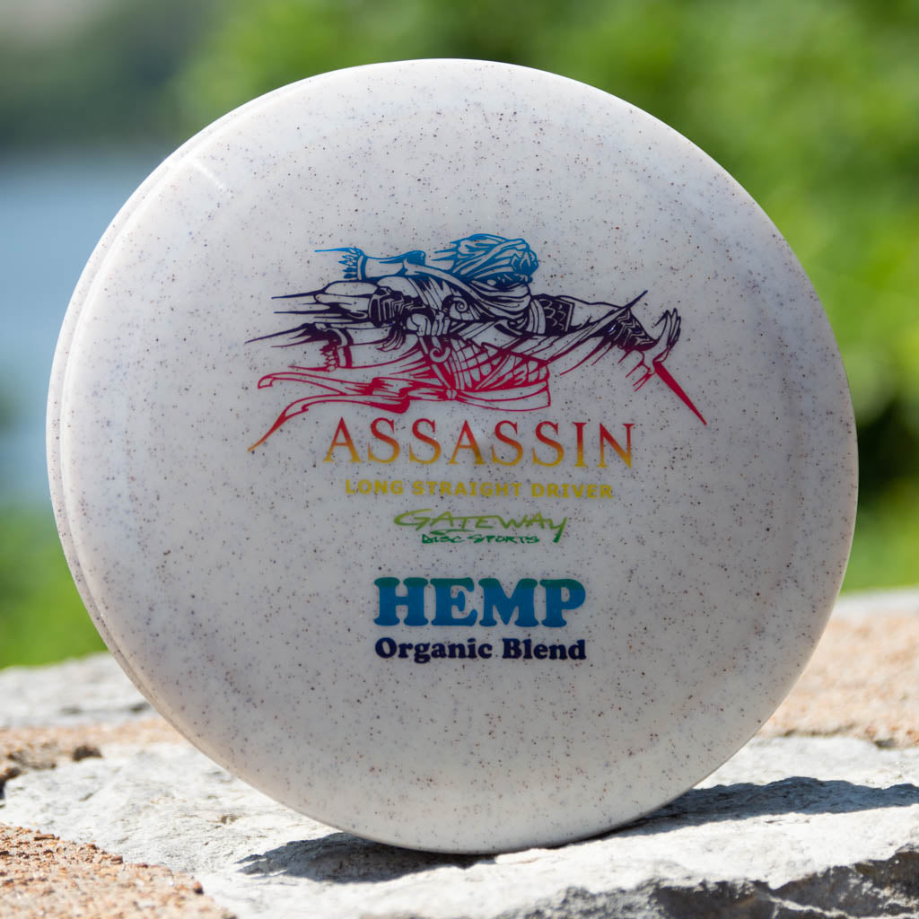 A white Assassin - Diamond Hemp distance driver with "neutral flight path" labeling rests on a rock outdoors, highlighting the quality craftsmanship of Gateway Disc Sports.